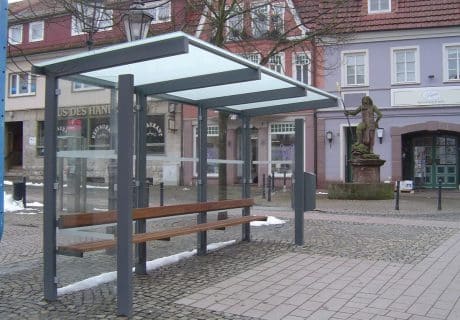 Eine graue dreifeldrige Wartehalle mit einer Holzbank über die gesamte Länge inklusive Anlehnholm